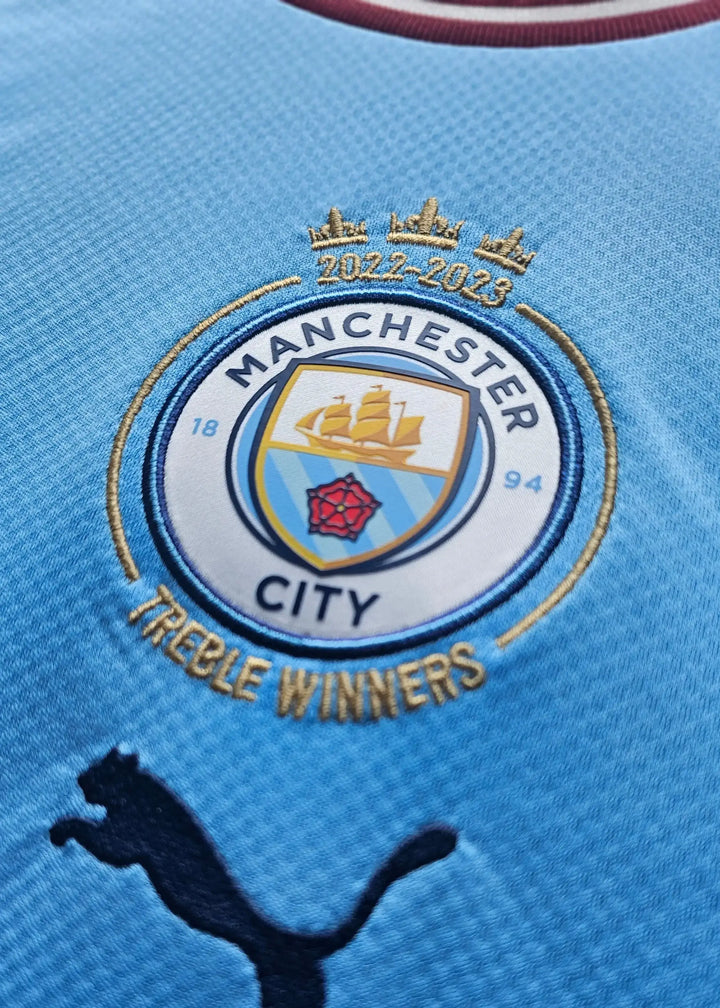 Close-up of Manchester City crest featuring Treble Winners text and 2022-23 year.