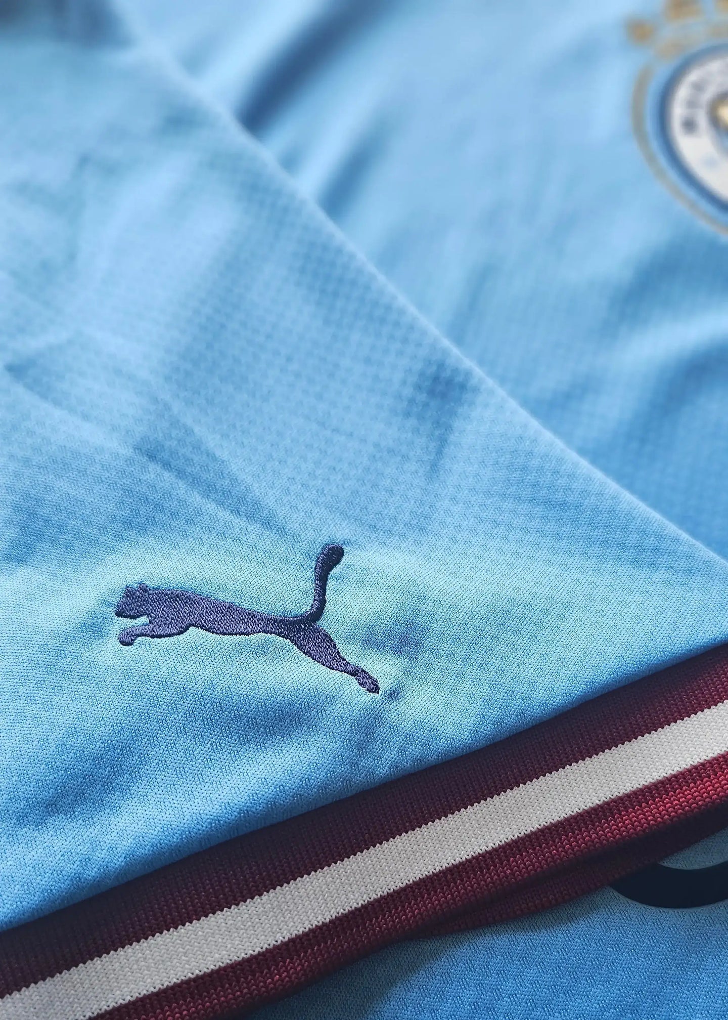 Detail view featuring the Puma logo on the light blue football shirt with maroon trim.