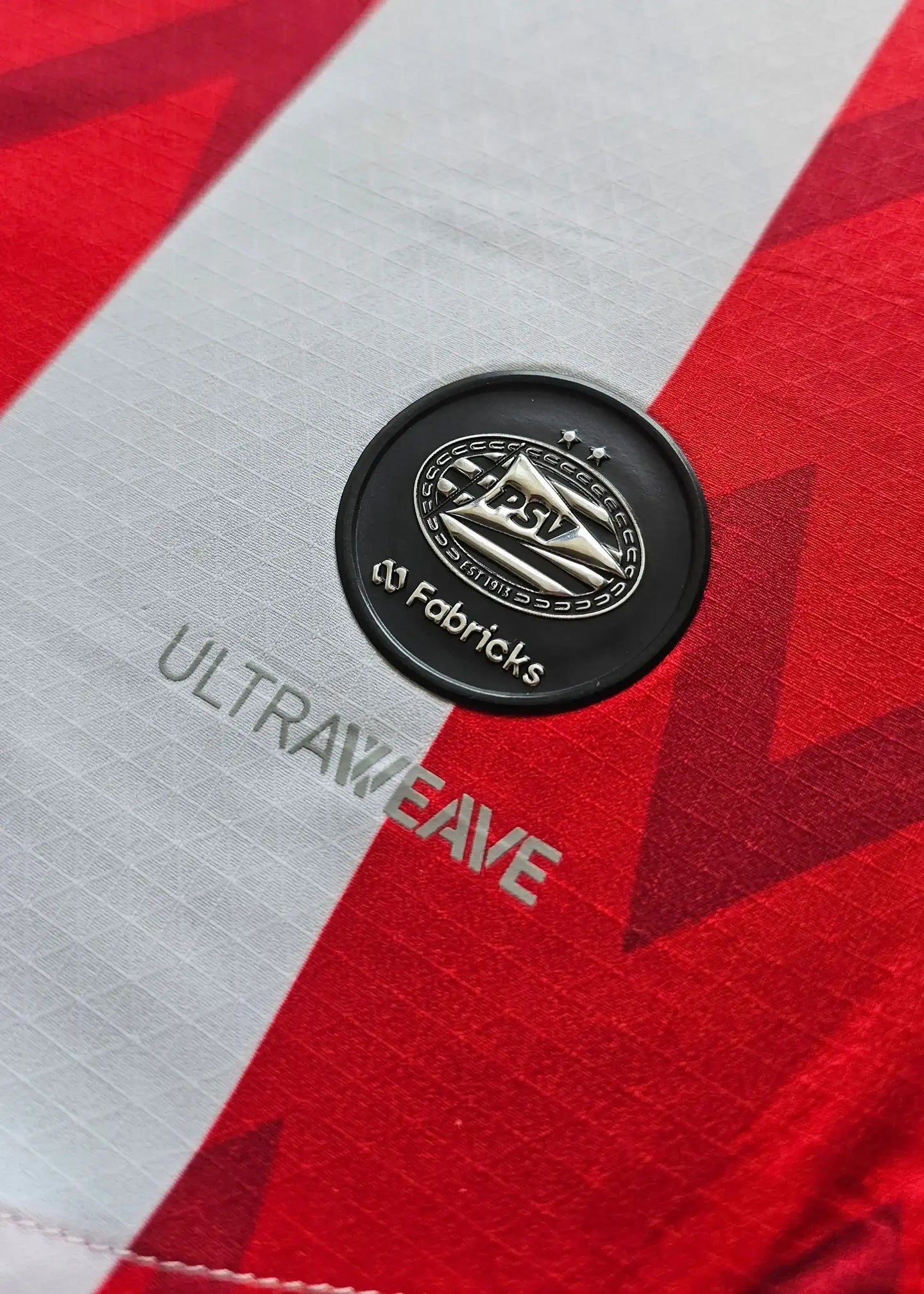 Close-up of PSV crest with two stars and 'ULTRAWEAVE' text on red and white striped background.