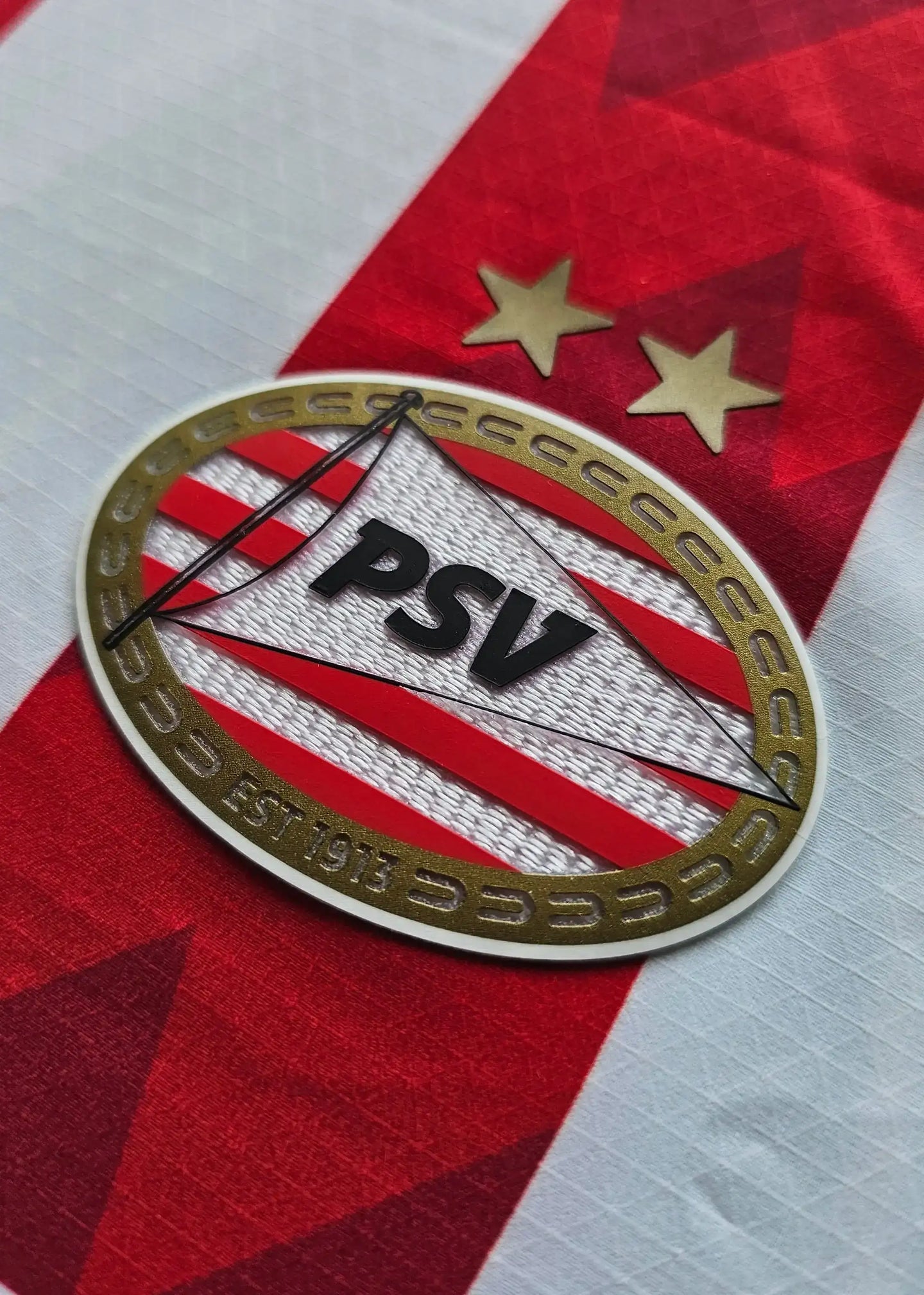 Close-up of PSV Eindhoven crest with two stars and established date 1913 on a red and white striped background.