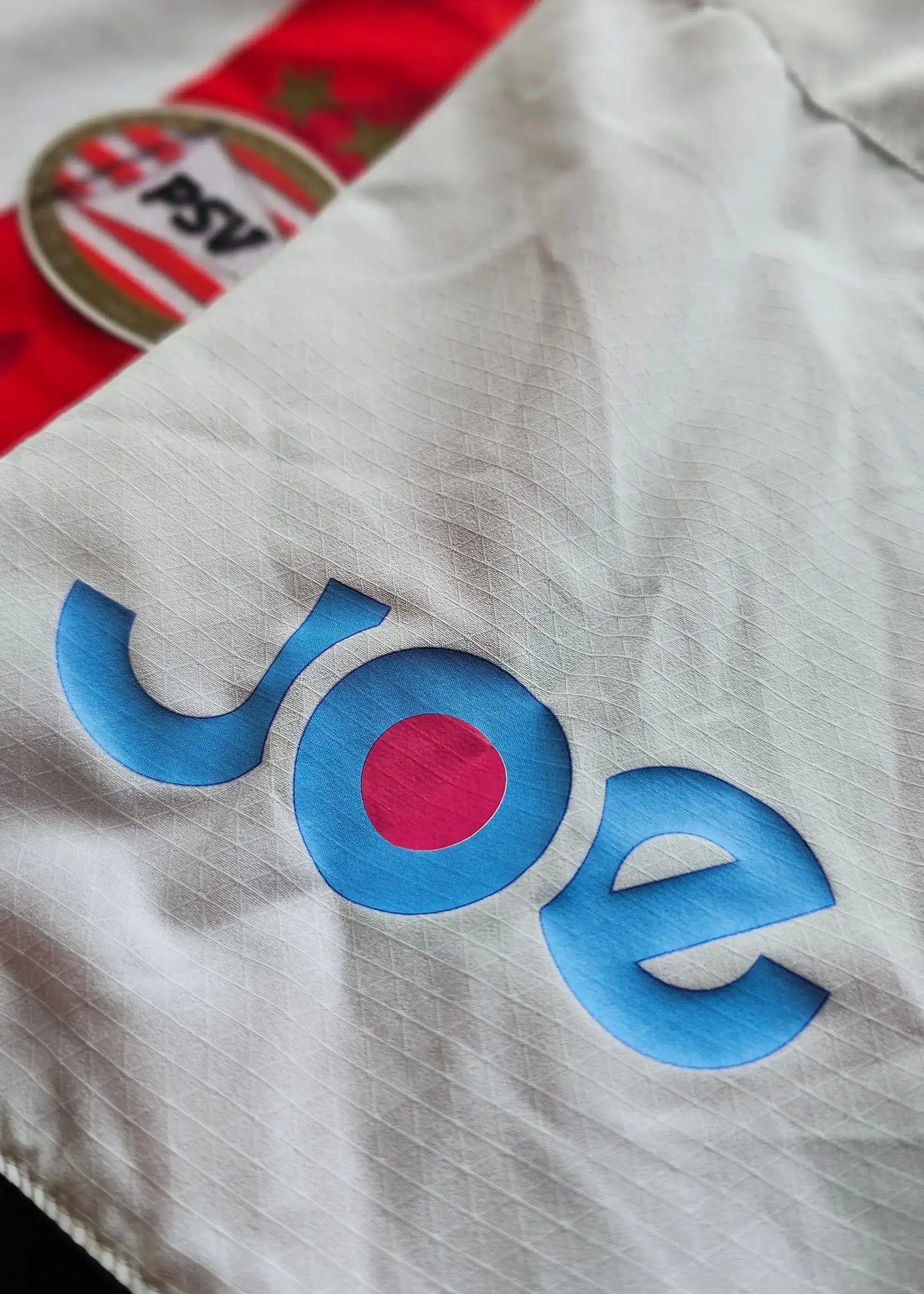 Detail showing the blue wordmark 'Joe' with a pink circle on a white football shirt.