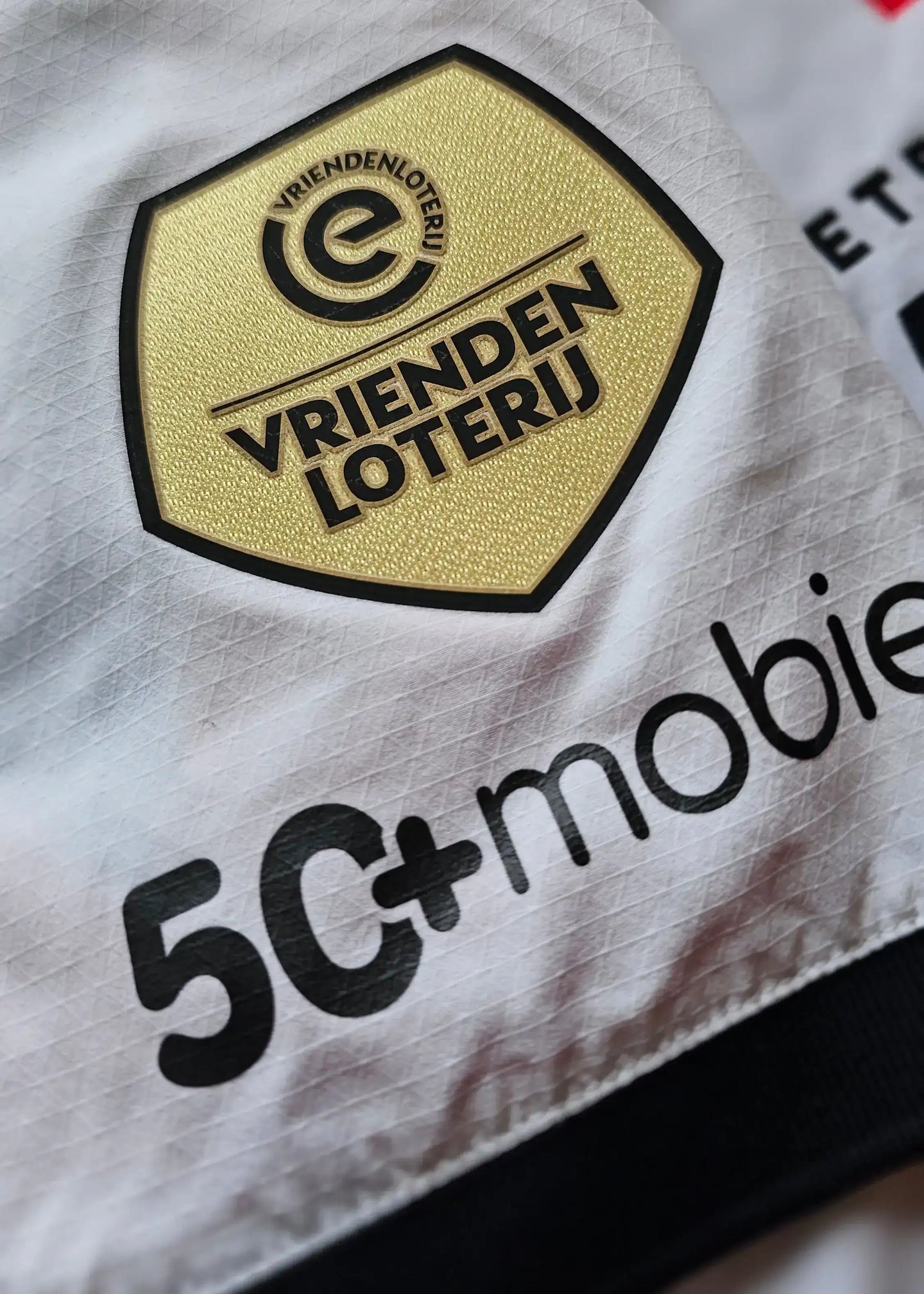 Gold and black VRIENDENLOTERIJ logo with 5C+mobile sponsor text on a white football shirt.
