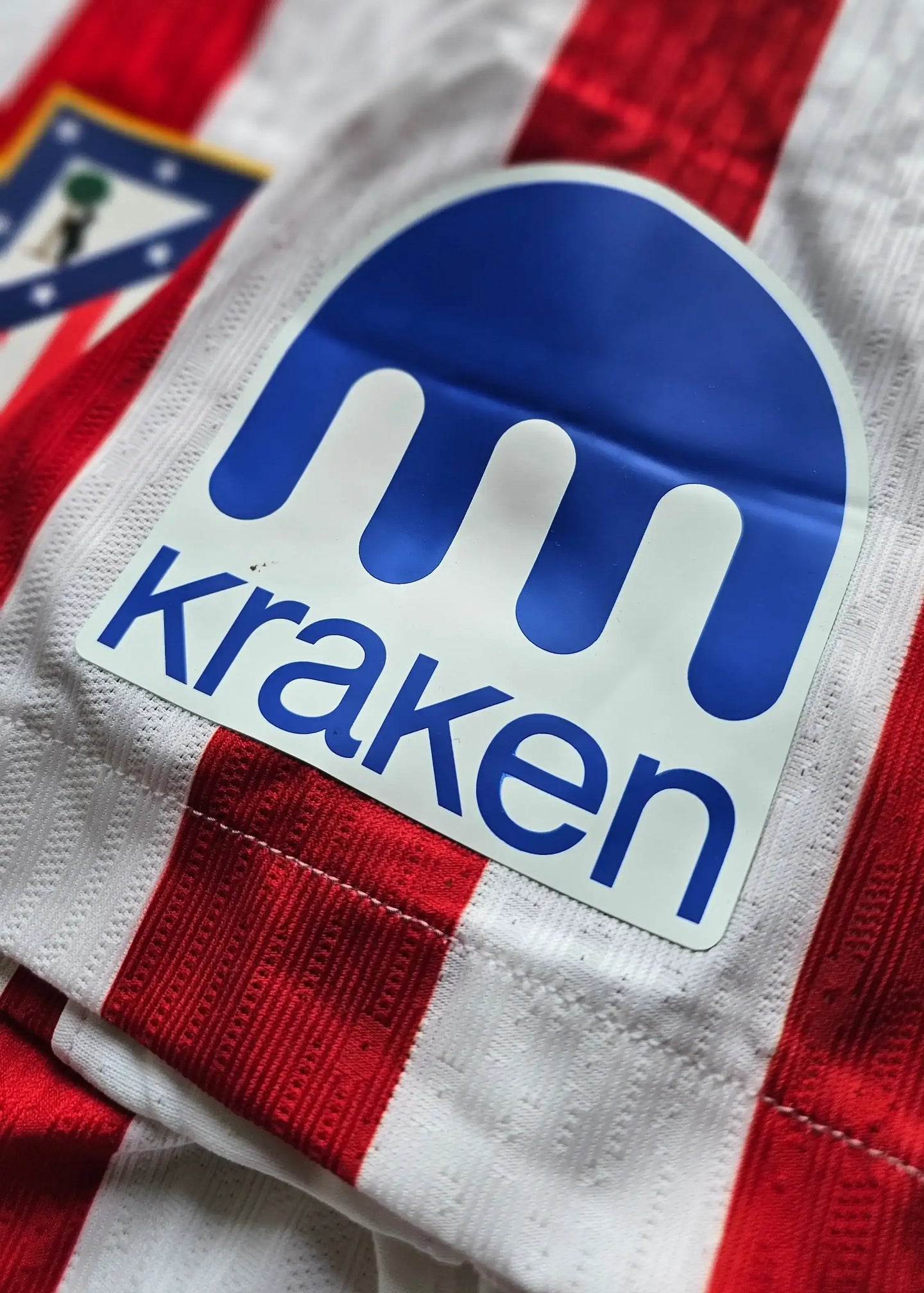 Close-up of Atlético Madrid crest and Kraken sponsor logo on a red and white striped football shirt.