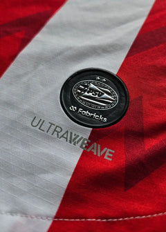PSV crest with two stars, ULTRAWEAVE fabric detail, and Fabricks label on red and white shirt.