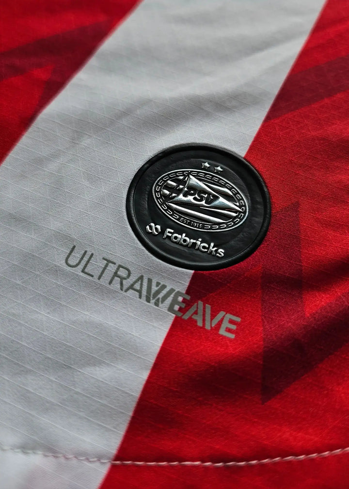 PSV crest with two stars, ULTRAWEAVE fabric detail, and Fabricks label on red and white shirt.