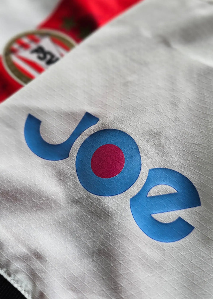 Close-up of PSV Eindhoven shirt featuring the blue 'Joe' logo and club crest, red and white stripes visible.