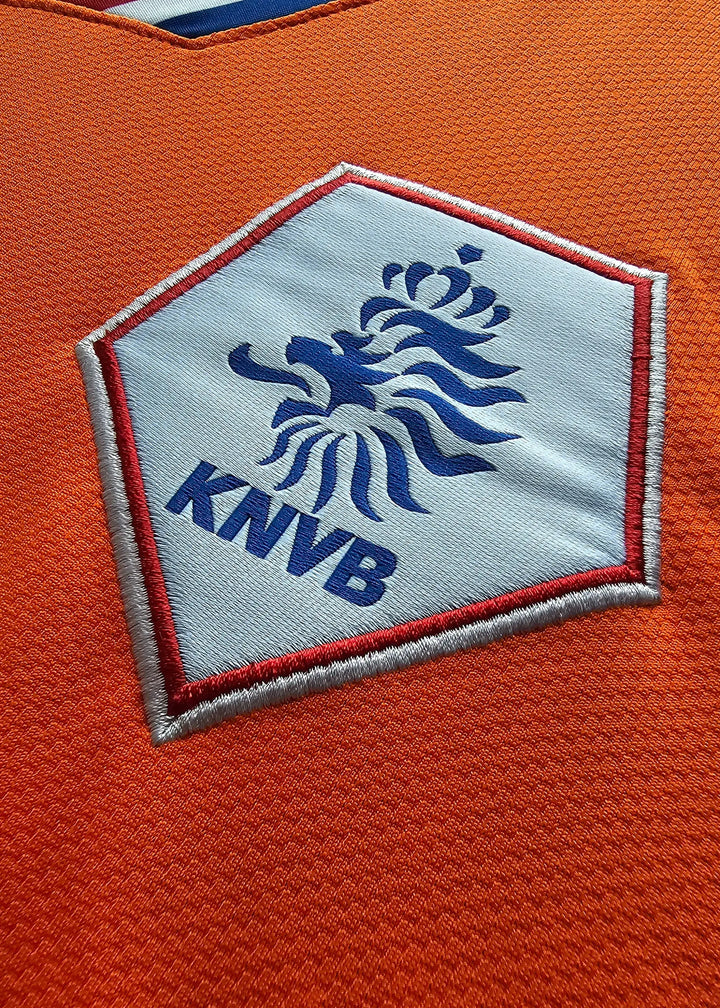 Close-up of the KNVB crest, its blue lion and crown embroidered on the pentagon patch with 'KNVB' text below, on Ruud van Nistelrooy's orange Holland shirt.