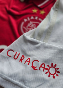 Front view showing Ajax crest with two stars and CURAÇAO printed in red on white fabric.