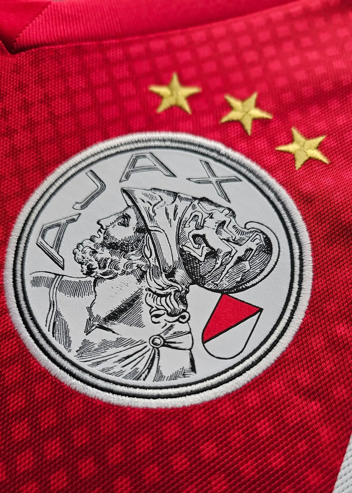 AFC Ajax crest on a red shirt with three gold stars above, featuring an embroidered design of a classical figure.
