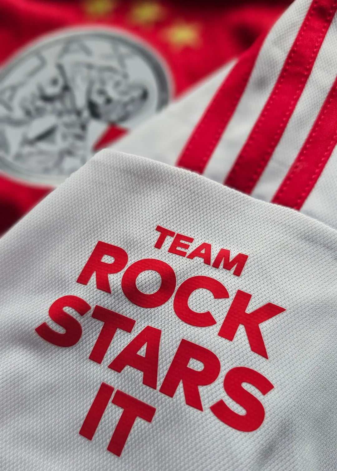 Detail of white fabric featuring the text TEAM ROCK STARS IT, with red stripes and Ajax crest visible.