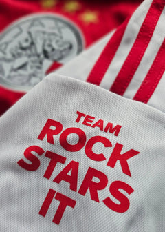 Detail of white fabric featuring the text TEAM ROCK STARS IT, with red stripes and Ajax crest visible.
