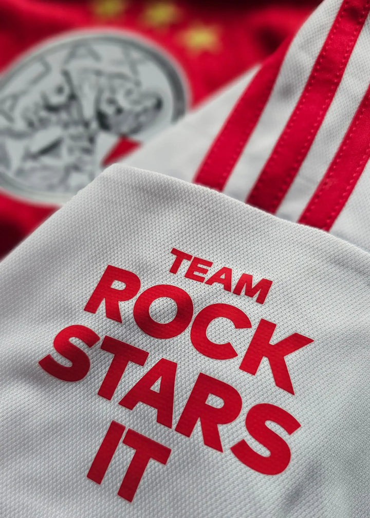 Detail of white fabric featuring the text TEAM ROCK STARS IT, with red stripes and Ajax crest visible.