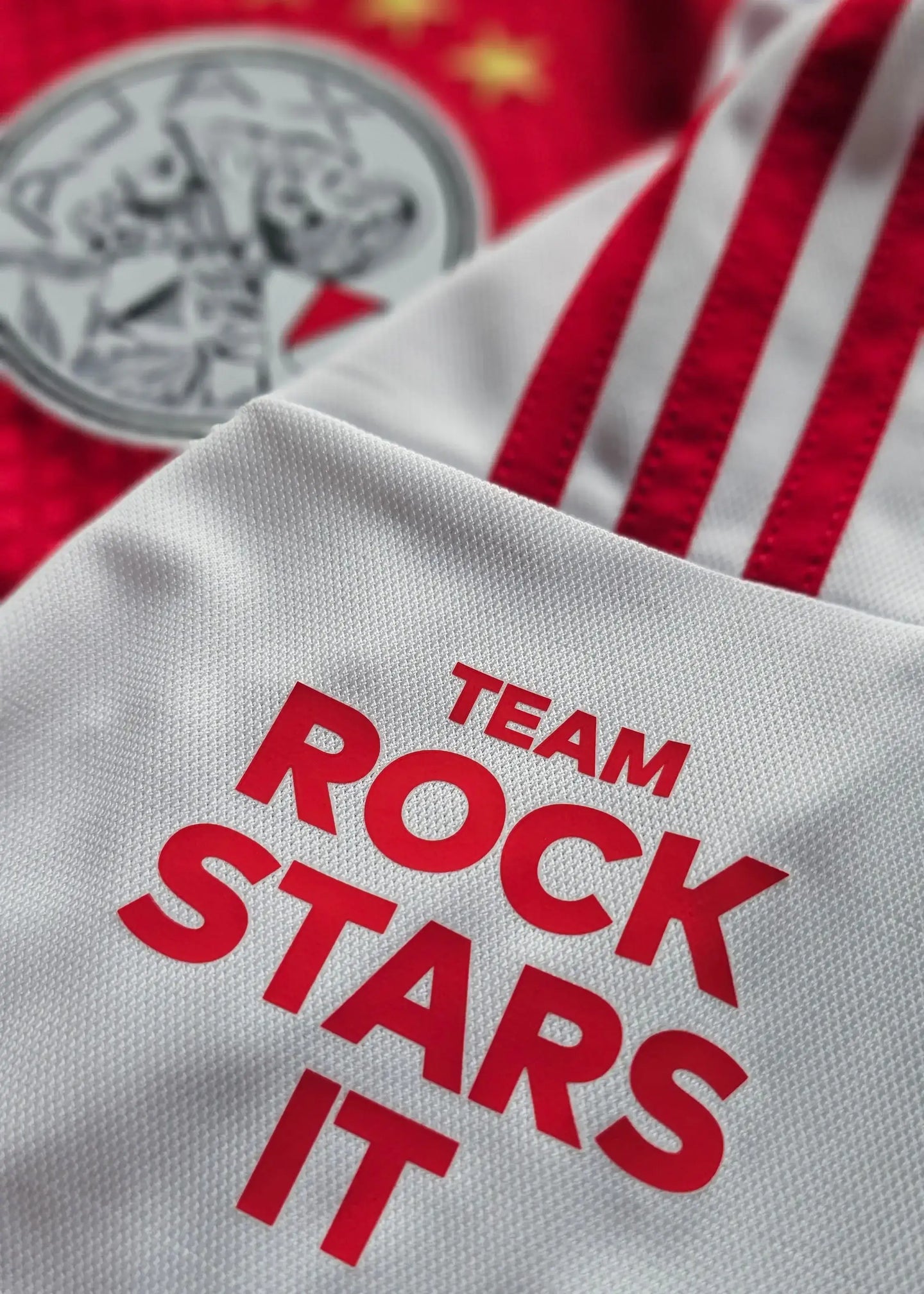 Close-up of AFC Ajax crest and white fabric with red 'TEAM ROCK STARS IT' text.