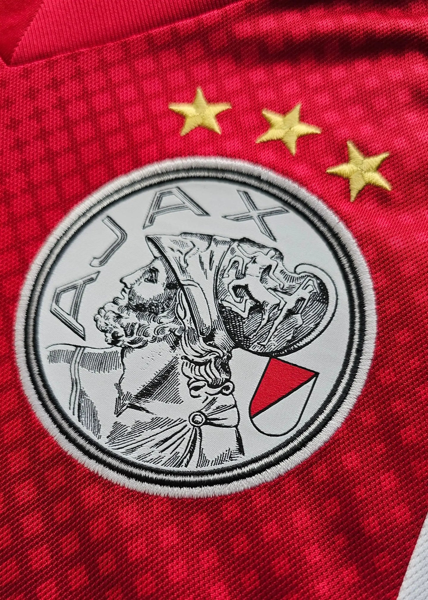 AFC Ajax crest with three gold stars above on a red background featuring detailed artwork.