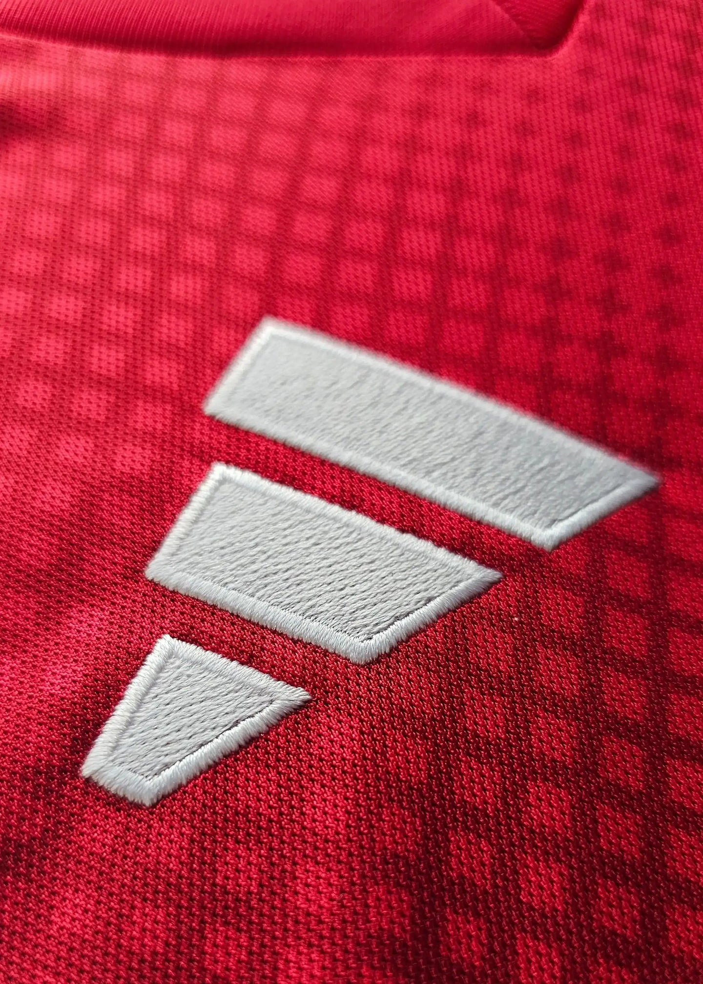 Close-up of the Adidas logo in white stitching on a red football shirt fabric.