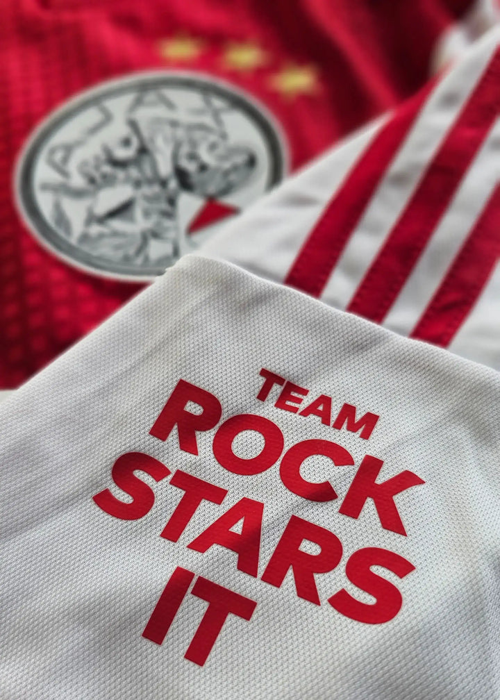 Detail showing Ajax crest with three stars and white fabric featuring 'TEAM ROCK STARS IT' in red.