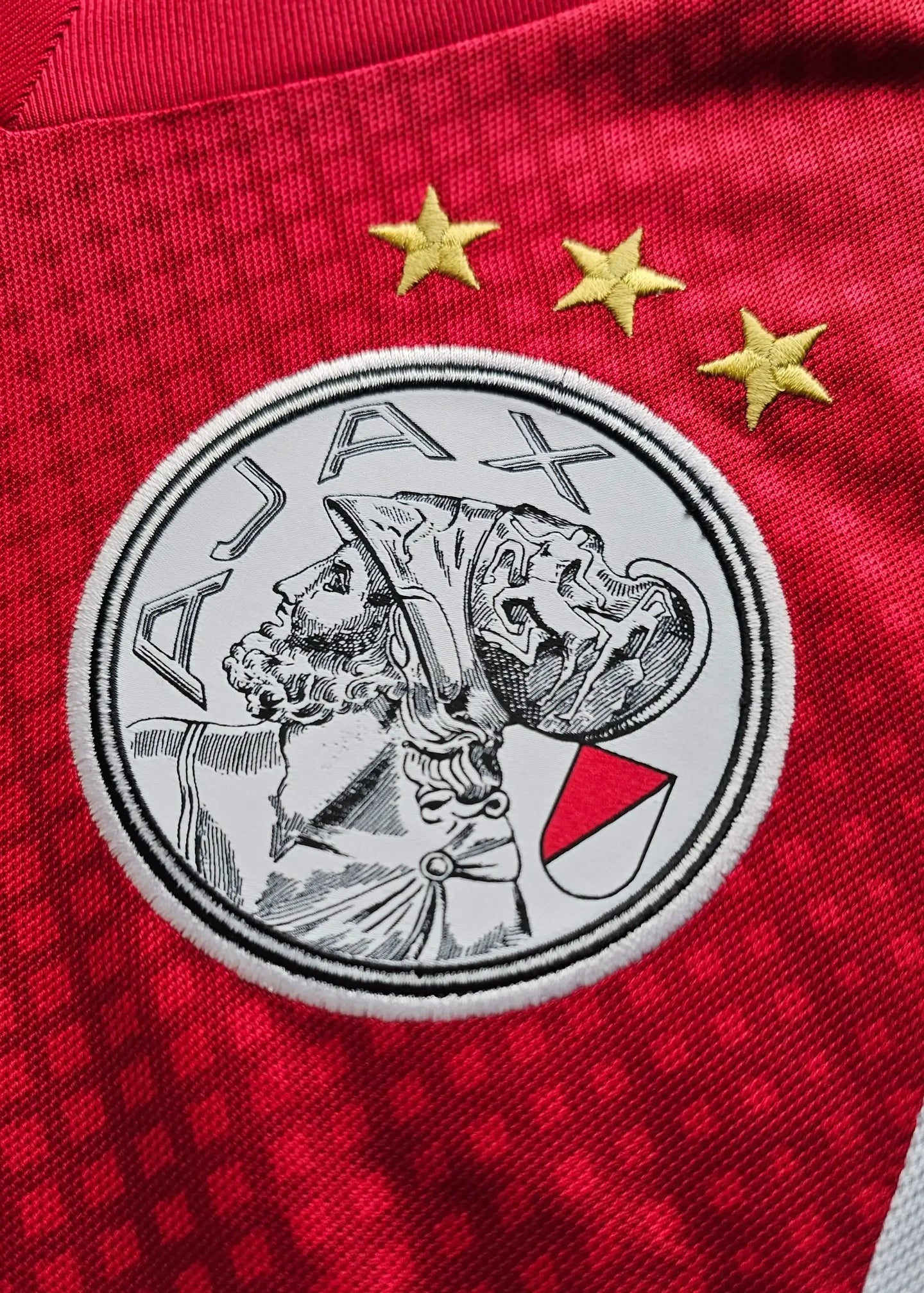 AFC Ajax crest with three gold stars, featuring a historical figure in profile, on a red jersey.