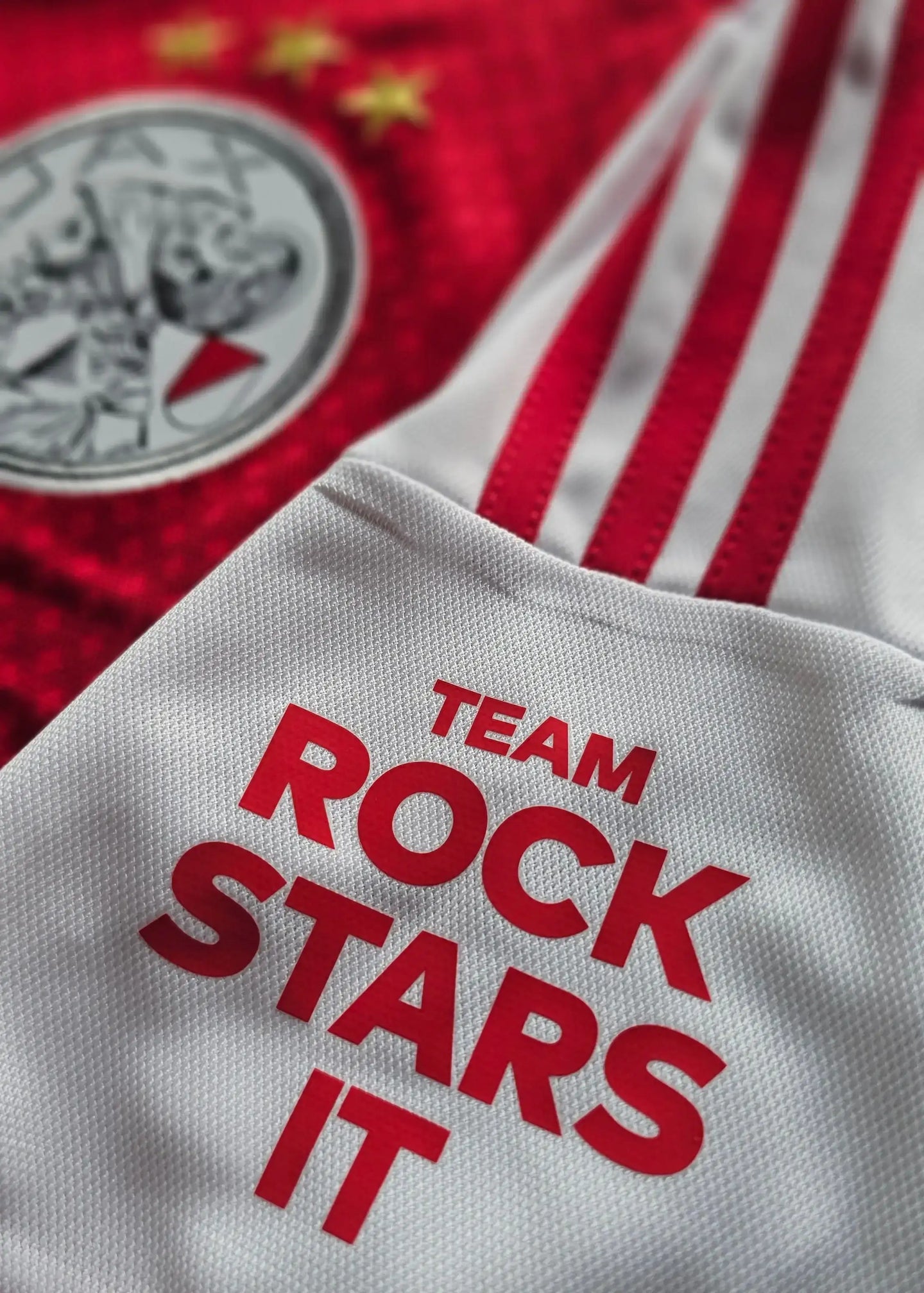Close-up of Ajax jersey featuring crest with three stars and white fabric with text 'TEAM ROCK STARS IT'.