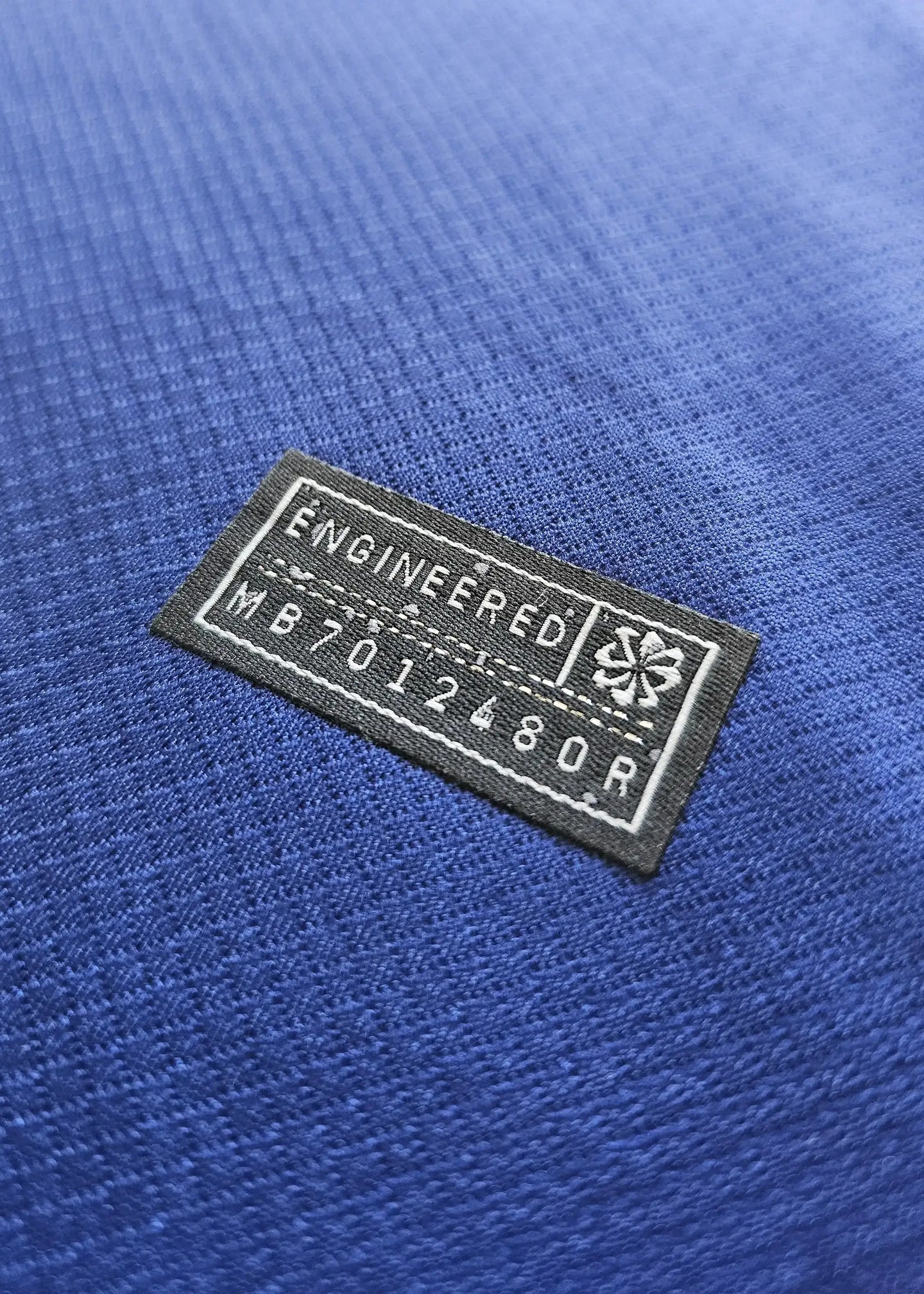 Close-up of the engineered label on a blue football shirt, showing text 'ENGINEERED' and 'MB 7012480R'.