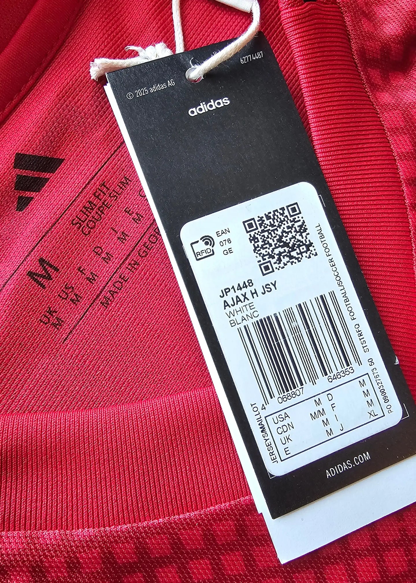 Close-up of size M label with adidas branding and AJAX H JSY details on a red football shirt.
