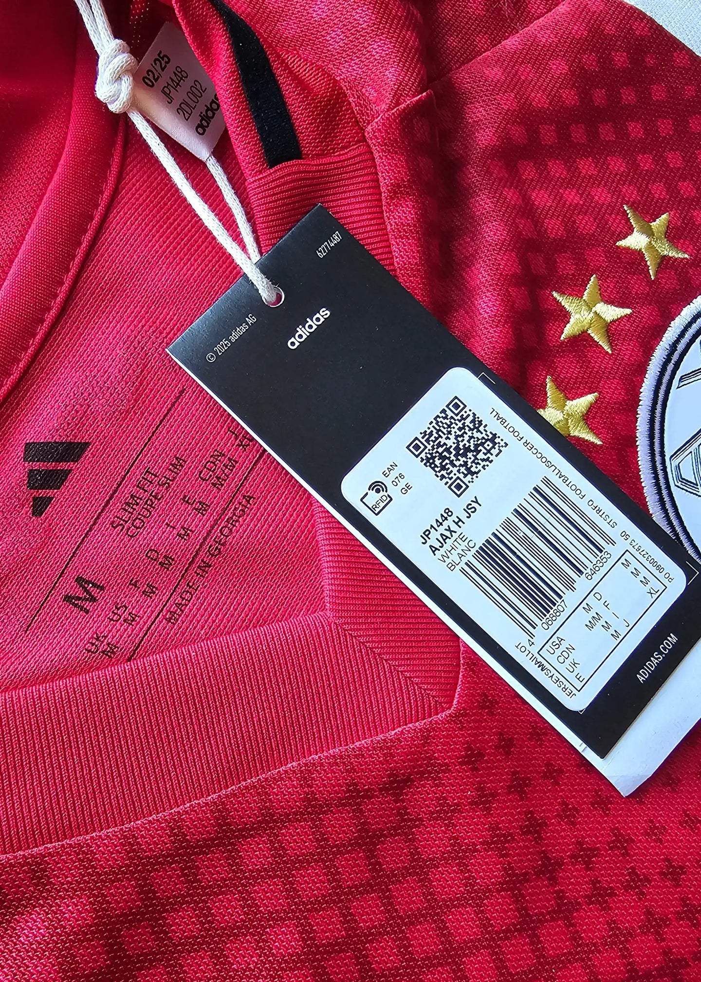 Close-up of the Adidas tag showing size 'M', red fabric with three gold stars and black detailing.