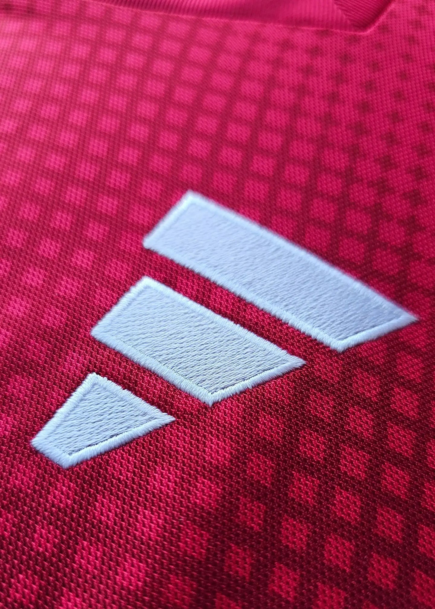 Close-up of the white adidas logo on the red textured fabric of the football shirt.