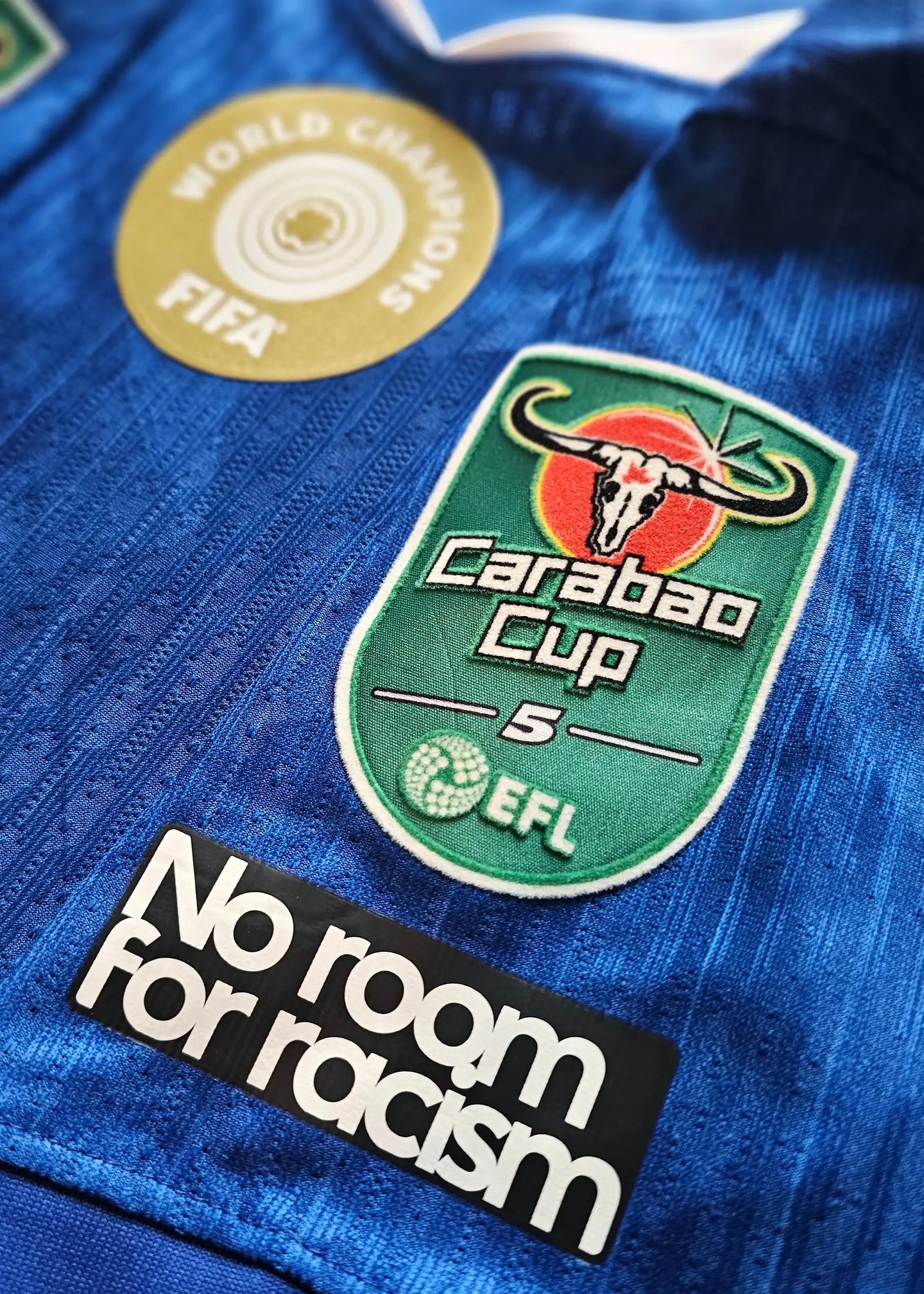 Close-up of Chelsea F.C. crest, Carabao Cup logo, and 'No room for racism' on a blue football shirt.