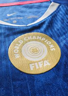 Crest detail with gold FIFA World Champions emblem on a blue football shirt.
