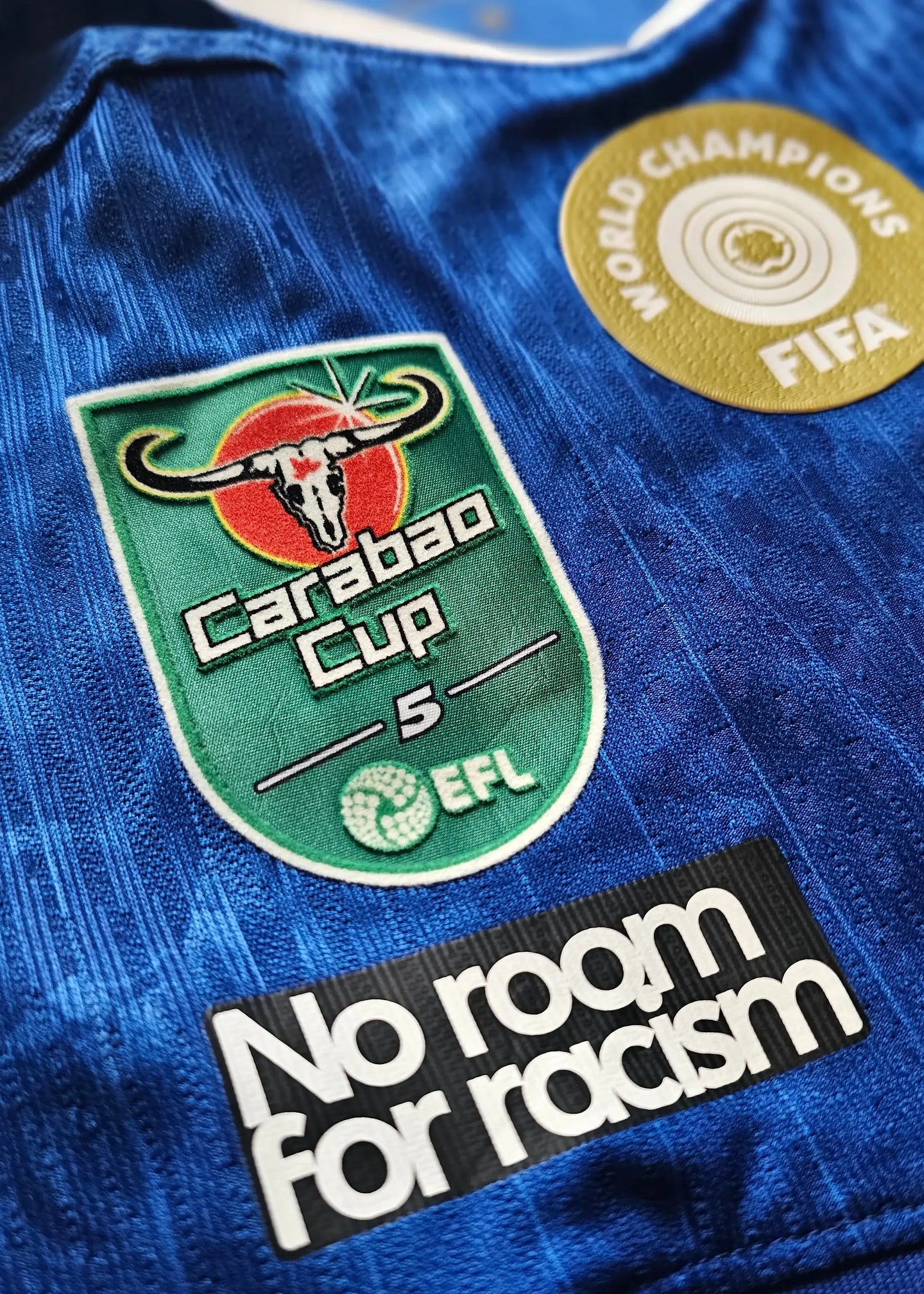 Close-up of Chelsea F.C. shirt showing Carabao Cup patch with 'No room for racism' text.