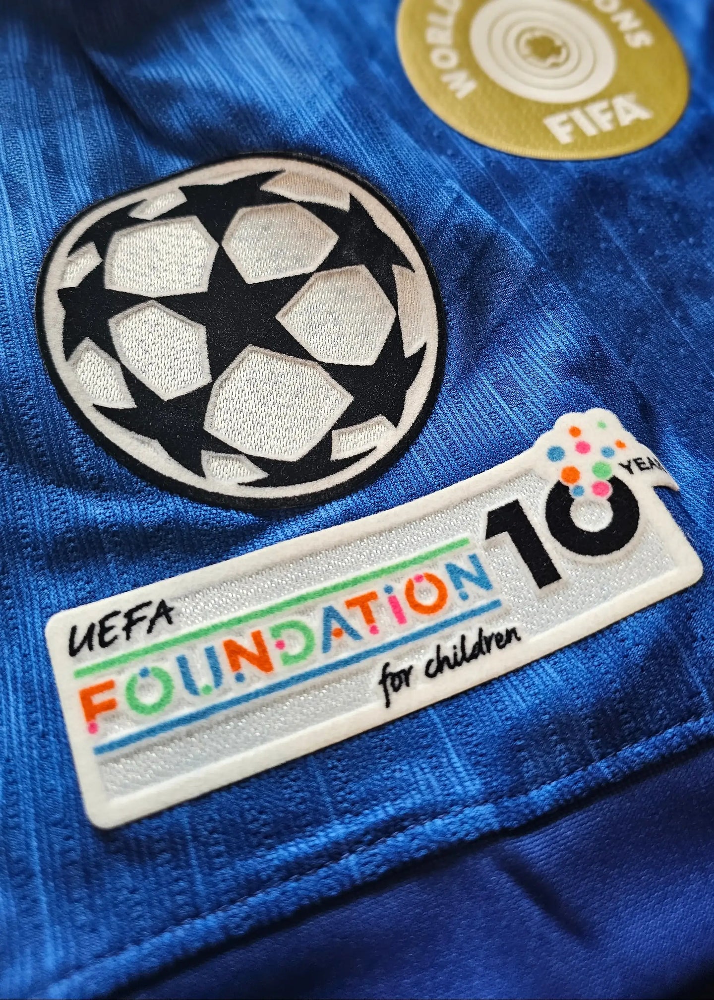 Close-up of UEFA Champions League logo and UEFA Foundation 10 years patch on a blue football shirt.