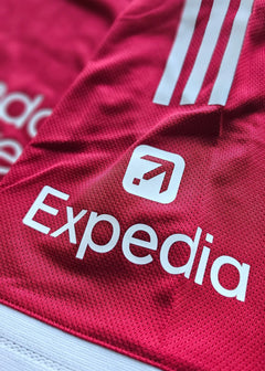 Close-up of the red football shirt featuring the Expedia logo and three white stripes.