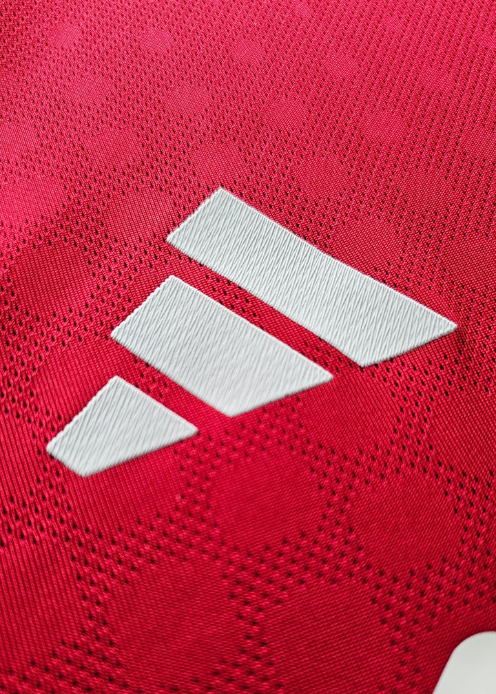 Close-up of white adidas logo on red football shirt fabric with textured hexagonal pattern.