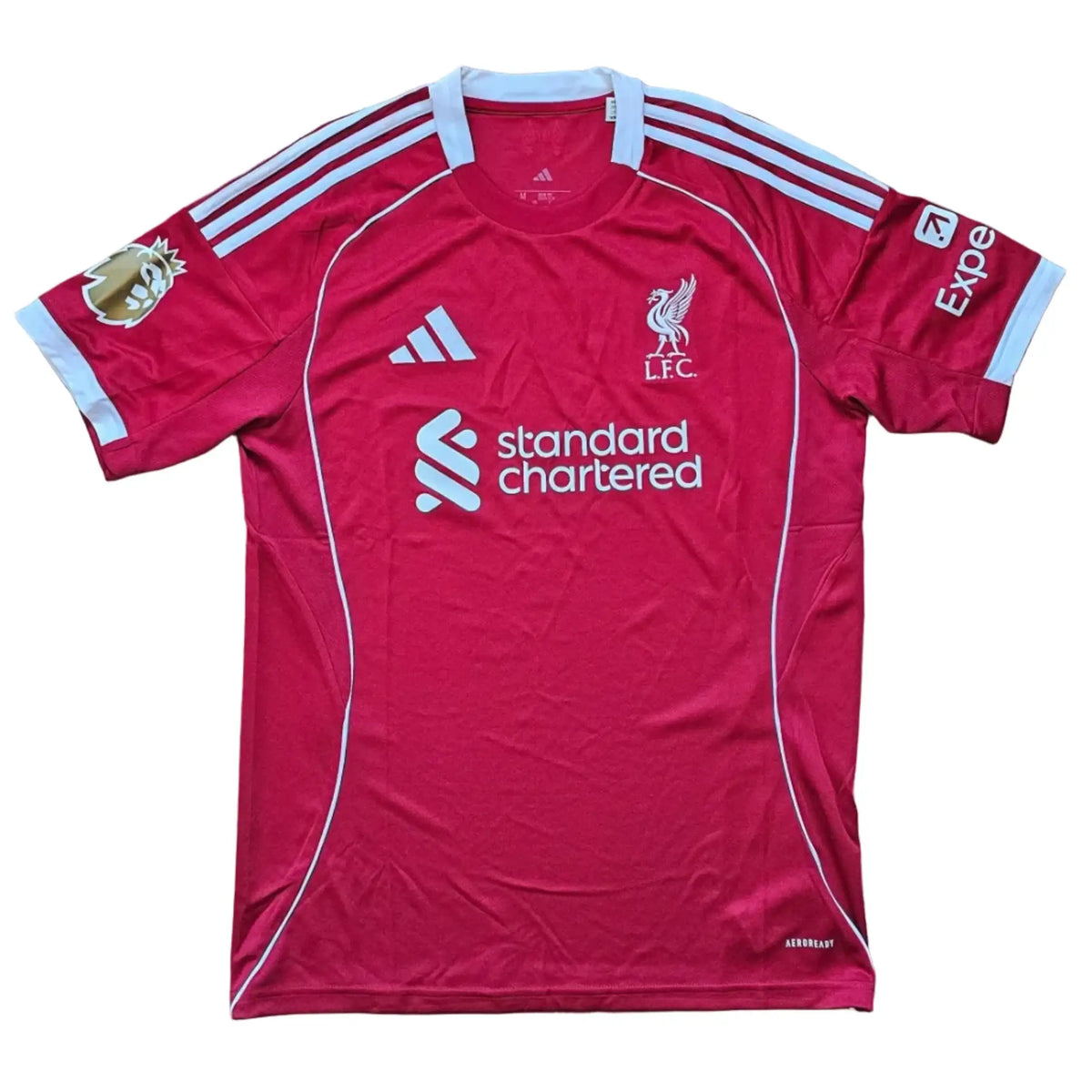Front of a red Liverpool F.C. 2025-2026 home shirt laid flat, adidas logo and club crest above the Standard Chartered sponsor, a gold-toned badge on one sleeve.