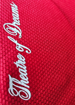 Close-up view of 'Theatre of Dreams' embroidered in white on red fabric.