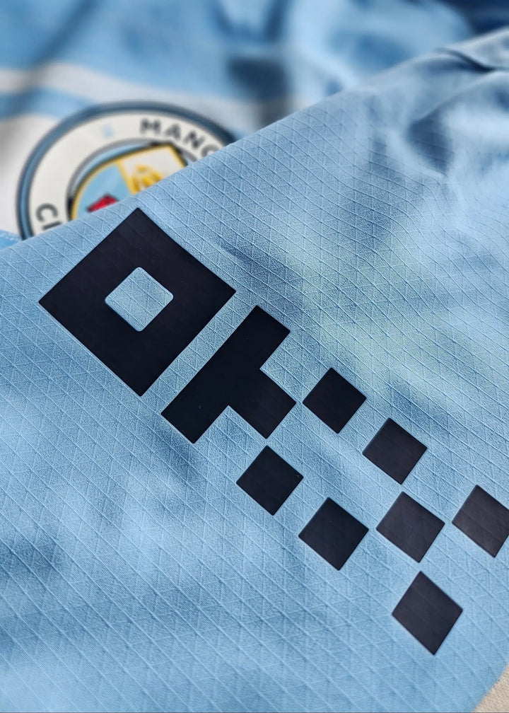 Close-up of Manchester City F.C. crest and dark blue sponsor logo on a light blue football shirt.