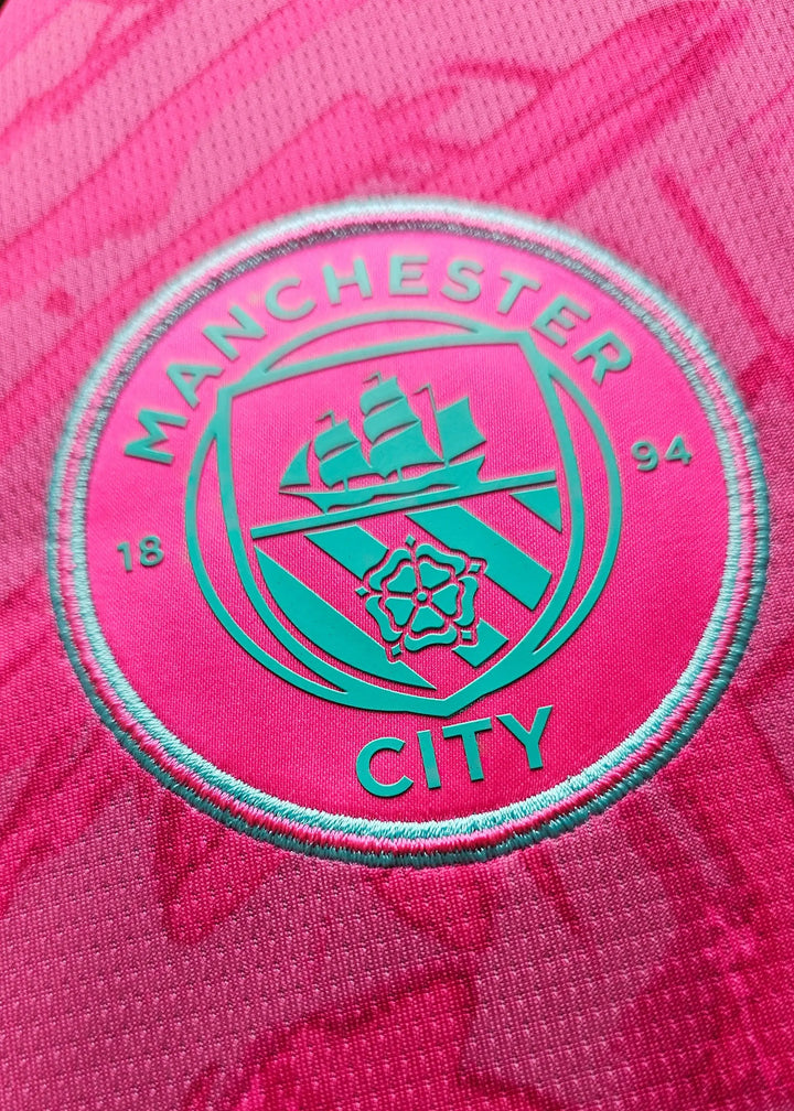 Crest of Manchester City with ship logo and 'MAN CITY' text on a pink background.