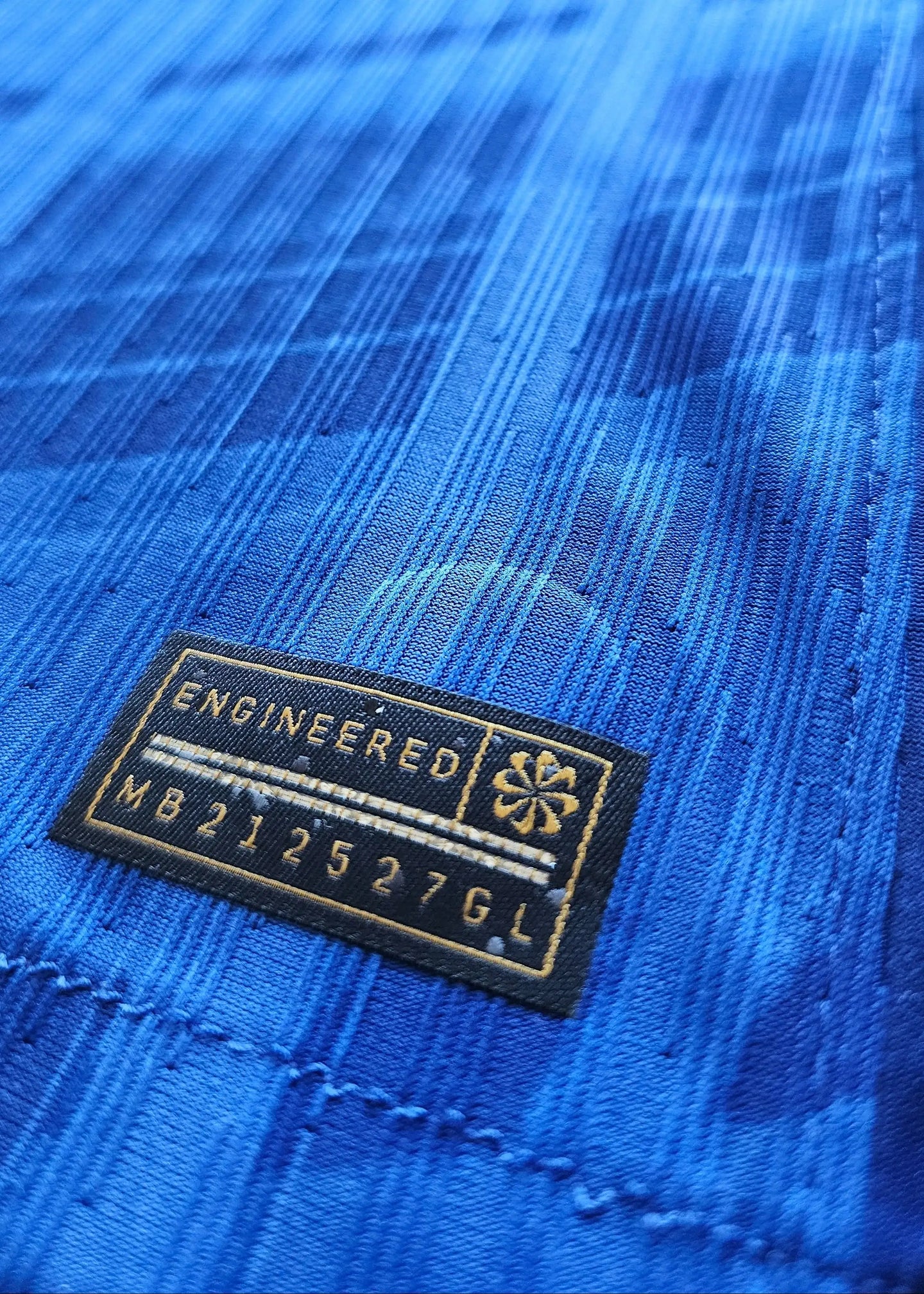 Close-up of the embroidered tag featuring 'ENGINEERED' and code 'MB212527GL' on a blue background.