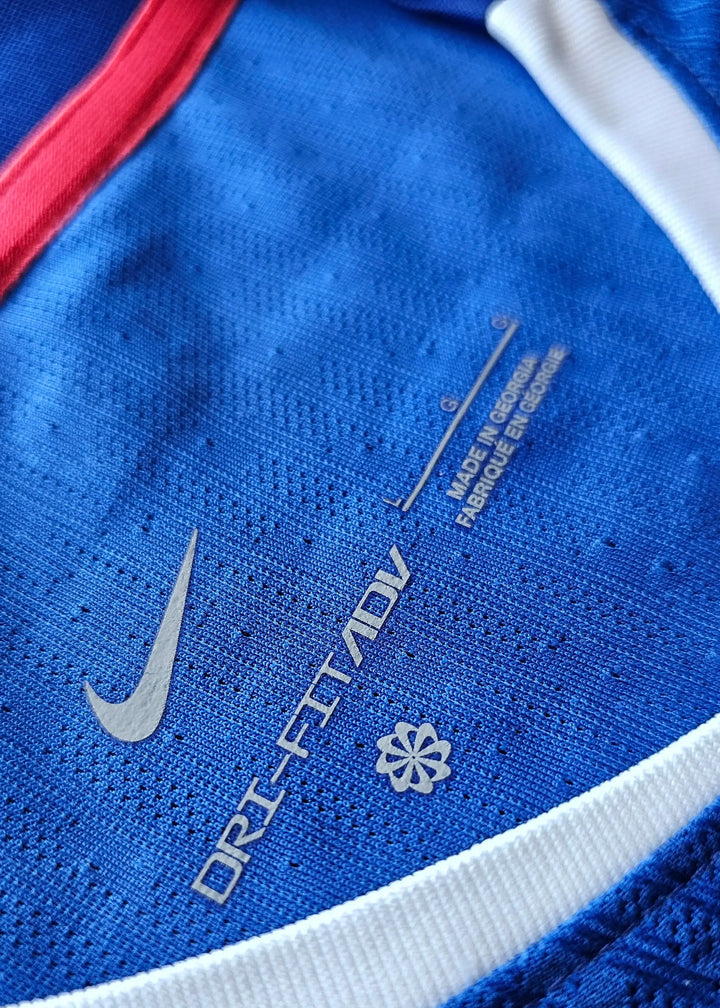 Close-up view of Nike DRI-FIT ADV label and logo on blue football shirt with red and white trim.