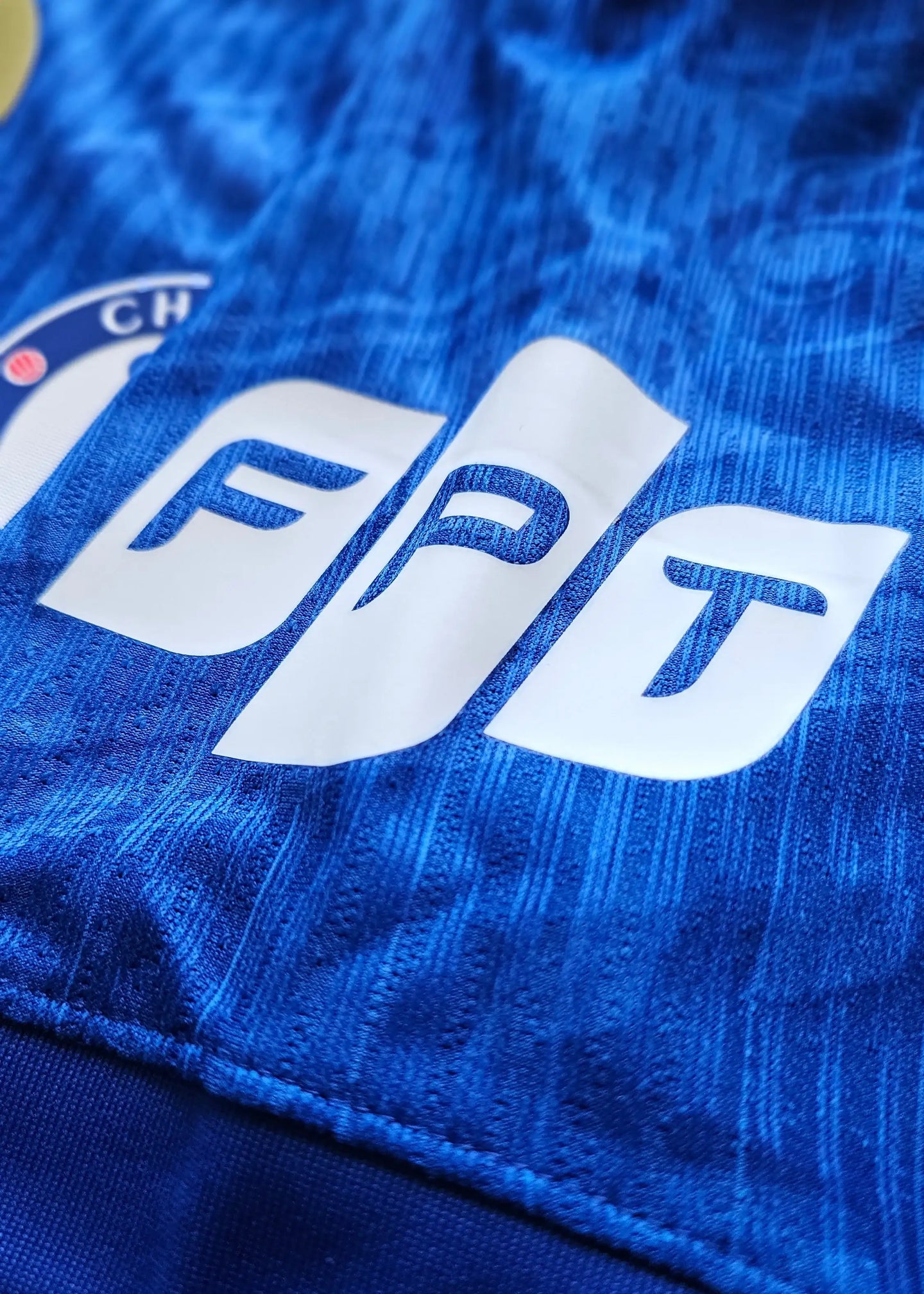 Close-up of Chelsea crest with FPT sponsor detail on a blue football shirt.