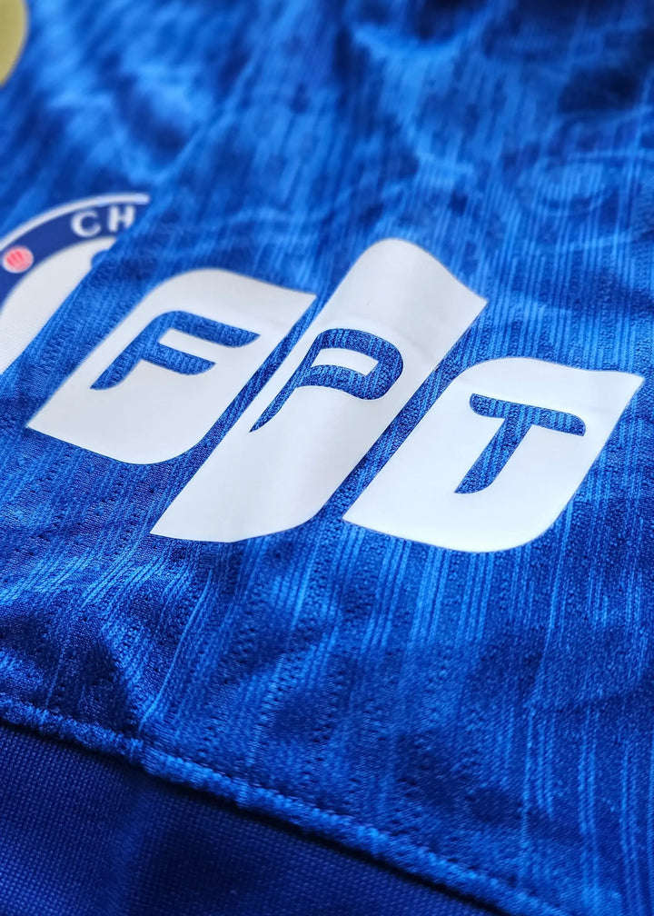 Close-up of Chelsea crest with FPT sponsor detail on a blue football shirt.
