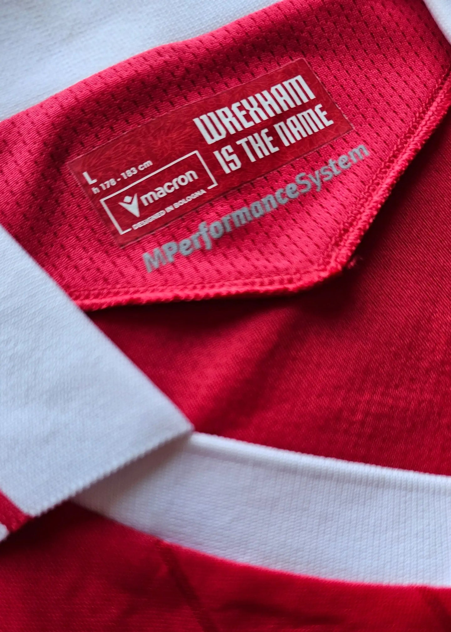 Crest detail showing 'WREXHAM IS THE NAME' label with Macron logo on red fabric.