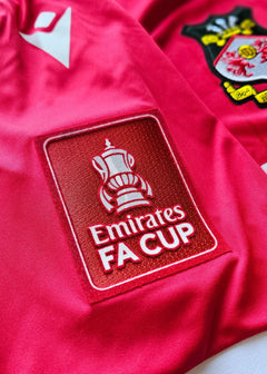 Detail of the Wrexham crest and Emirates FA Cup patch on a red football shirt.