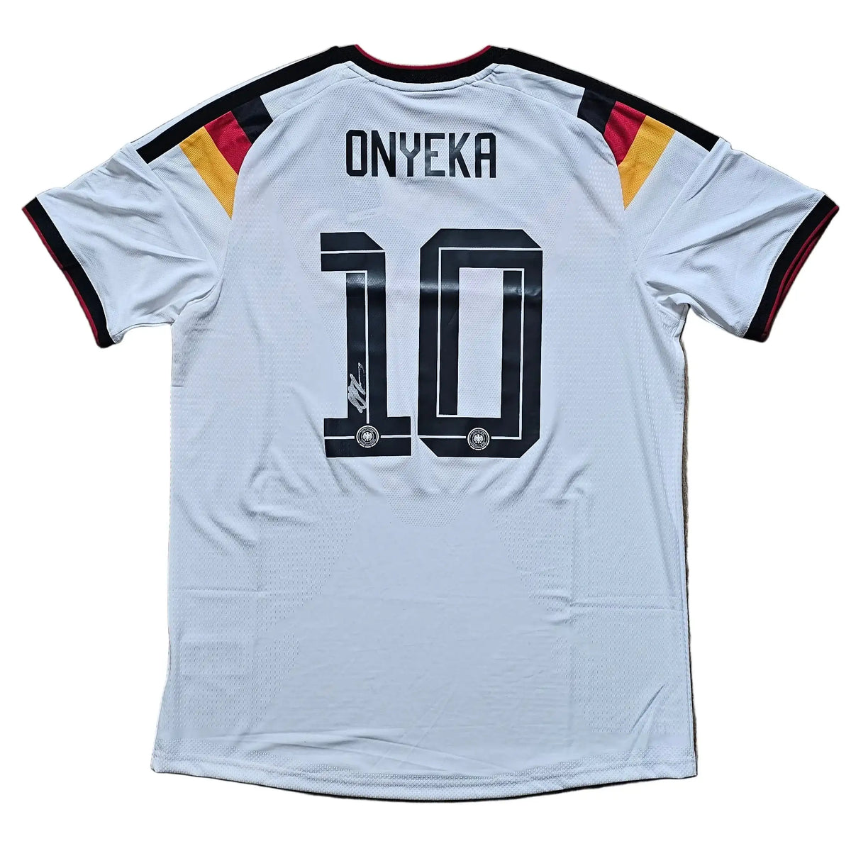A white Germany national team soccer jersey with 'ONYEKA' and the number '10' printed on the back, laid flat on a light brown wooden floor. The jersey features black trim on the collar and sleeve cuffs, with black, red, and gold stripes on the shoulders.
