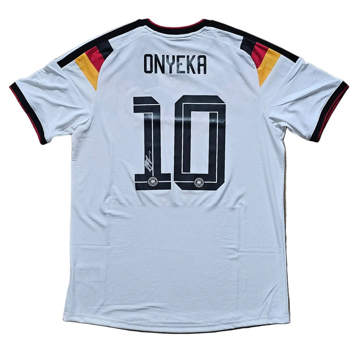 A white Germany national team soccer jersey with 'ONYEKA' and the number '10' printed on the back, laid flat on a light brown wooden floor. The jersey features black trim on the collar and sleeve cuffs, with black, red, and gold stripes on the shoulders.