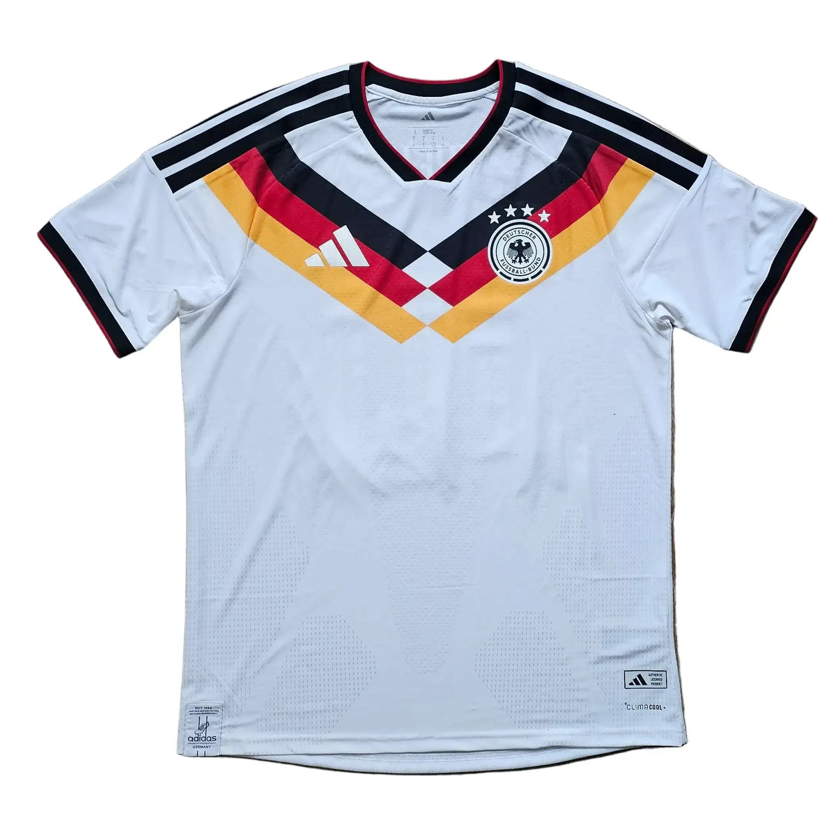 A white Germany national team soccer jersey by Adidas, featuring the black, red, and gold V-neck design and three stars on the chest, laid flat on a light brown wooden floor.