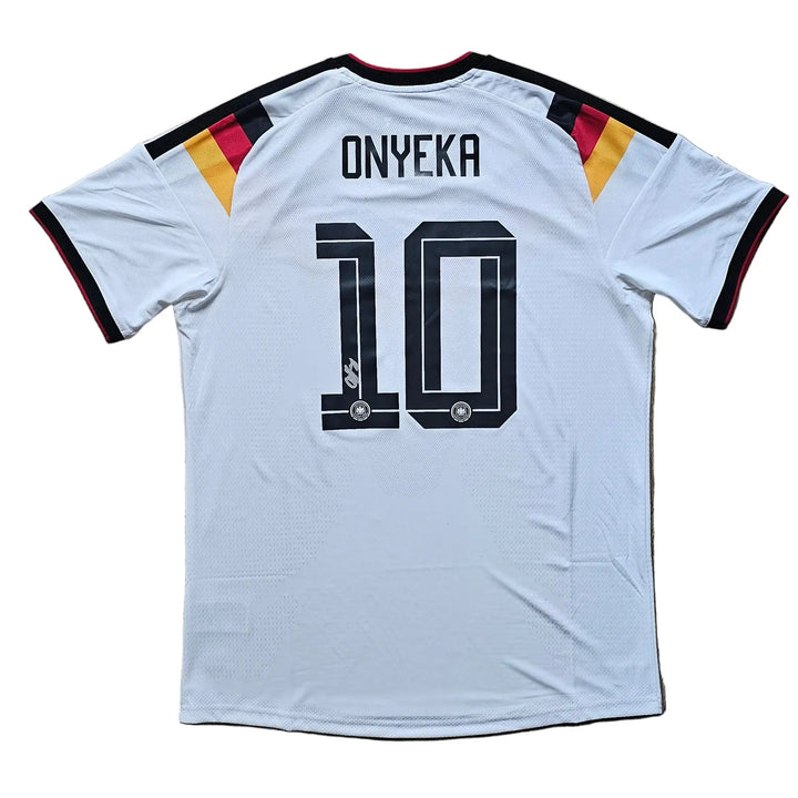 A white Germany national team soccer jersey with 'ONYEKA' and the number '10' printed on the back, laid flat on a light brown wooden floor. The jersey features black trim on the collar and sleeve cuffs, with black, red, and gold stripes on the shoulders.