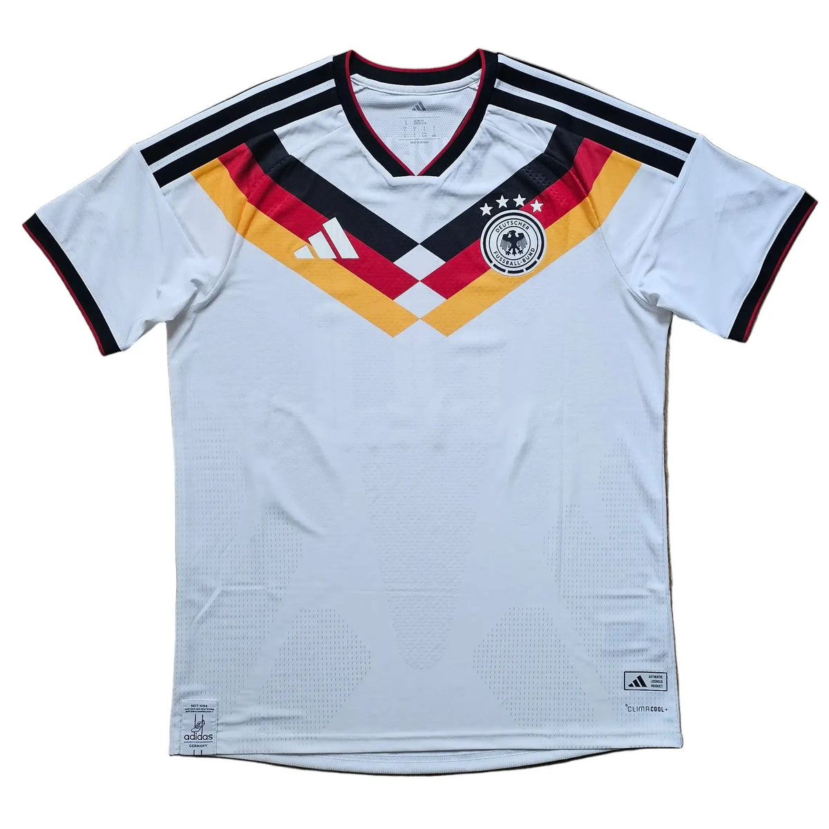 A white Germany national team soccer jersey by Adidas, featuring the German flag chevron on the chest and black, red, and gold trim, laid flat on a light brown wooden floor.