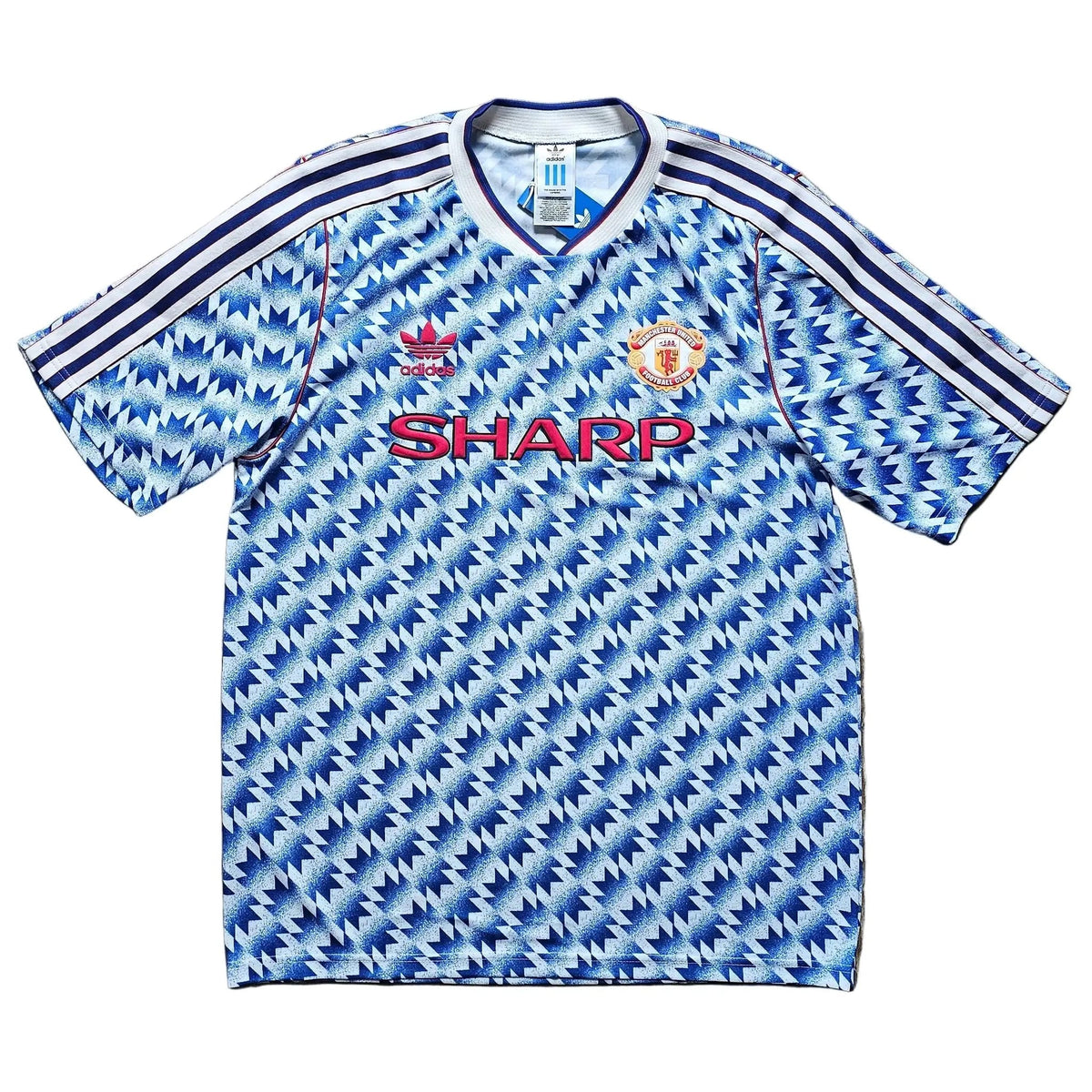 A vintage Manchester United short-sleeve soccer jersey with a blue and white geometric pattern, laid flat on a wooden floor. It features the red 'SHARP' sponsor logo on the chest and the Manchester United crest on the right side.