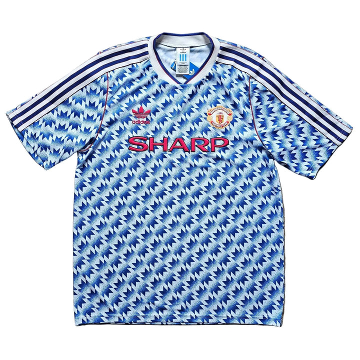 A vintage Manchester United short-sleeve soccer jersey with a blue and white geometric pattern, laid flat on a wooden floor. It features the red 'SHARP' sponsor logo on the chest and the Manchester United crest on the right side.