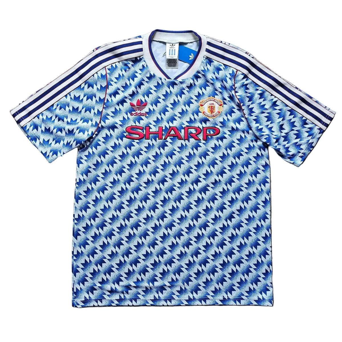 A vintage Manchester United short-sleeve soccer jersey with a blue and white geometric pattern, featuring the red 'SHARP' sponsor logo on the chest and the club crest on the right side. The jersey is laid flat on a light brown wooden floor.